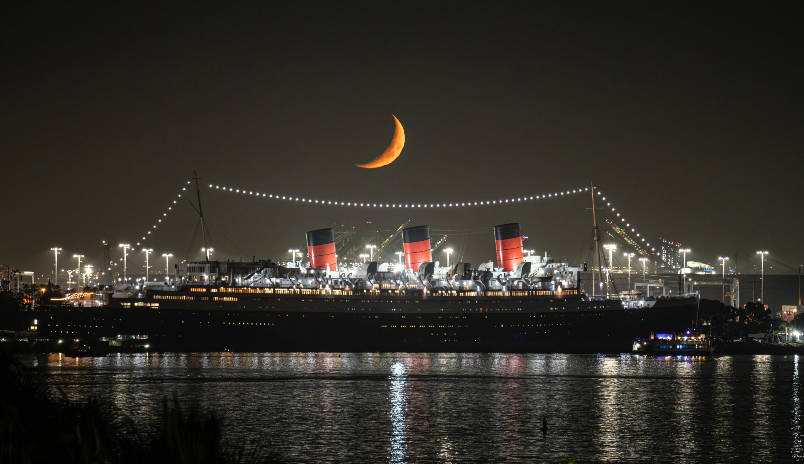 The Queen Mary ship illuminated at night under a crescent moon in Long Beach harbor.